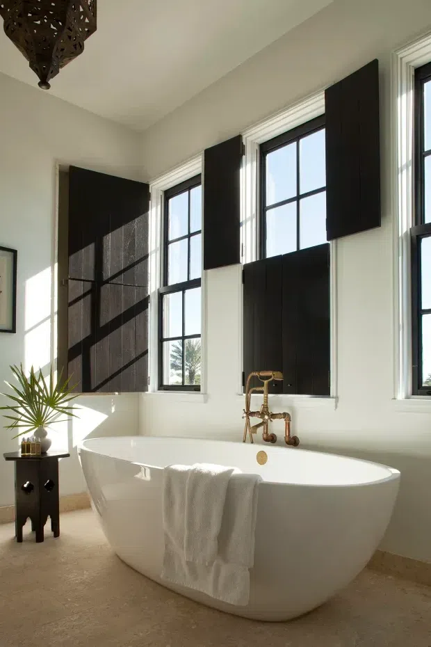 Bright, elegant bathroom featuring a freestanding white bathtub beneath tall black-framed windows. The room has pale walls, warm beige stone or tile flooring, dark timber window shutters/panels, and an exposed brass wall-mounted bath tap. A small dark side table with a potted plant and a decorative hanging pendant add character.