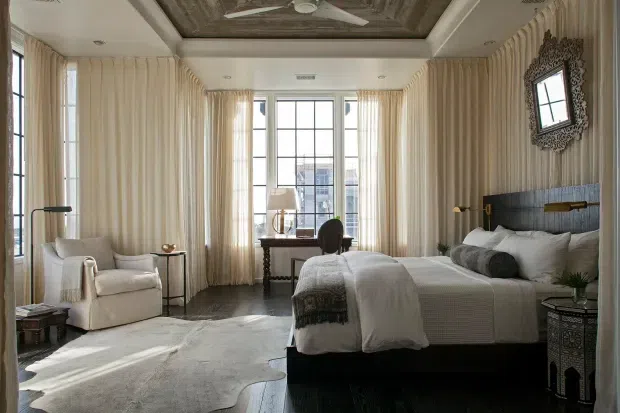 A refined bedroom with two beds, a dark upholstered headboard, and floor-to-ceiling cream curtains framing tall divided-pane windows. The room includes a writing desk by the window, an upholstered armchair, bedside tables, layered bedding, and a large pale rug. A decorative dark framed mirror hangs on the paneled wall, while the ceiling features a dark inset decorative panel with a ceiling fan. The palette is warm neutral with dark contrasting furniture and a calm, luxurious hotel-like atmosphere.