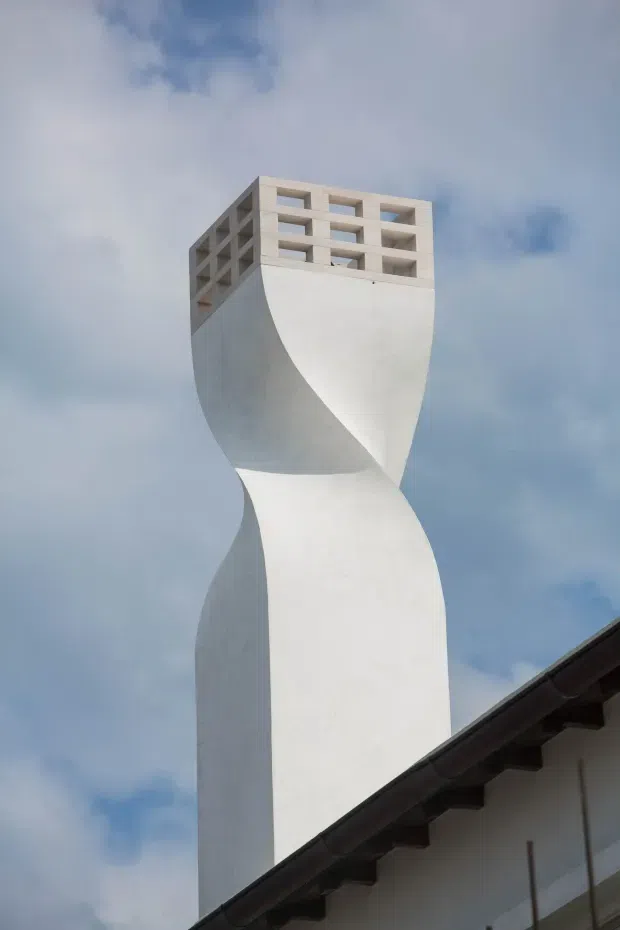 A dramatic close-up exterior view of a tall white sculptural architectural tower or chimney, formed with sweeping curved concrete or plastered surfaces that twist inward at the centre. The upper section has a rectangular open-brick or concrete-block screen with repeated horizontal openings. A pitched dark roof from an adjoining building crosses the lower foreground, beneath a partly cloudy blue sky.