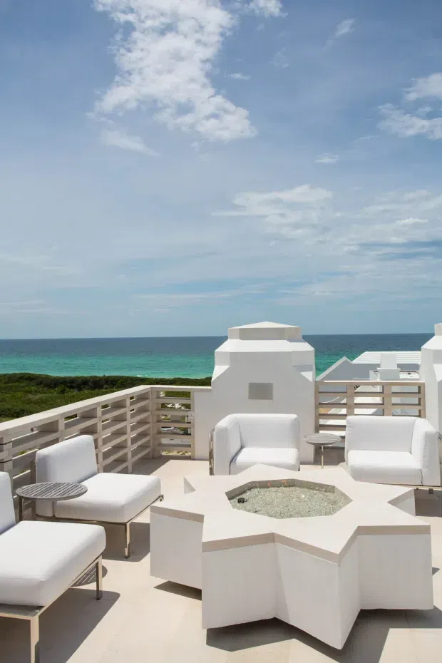 A bright rooftop or elevated outdoor terrace overlooking the ocean, furnished with white cushioned lounge chairs and a large white geometric built-in fire table or central feature. White parapet walls and horizontal privacy railings frame the coastal view, with low vegetation between the terrace and turquoise water. The scene has a clean, resort-like contemporary aesthetic under a blue sky.
