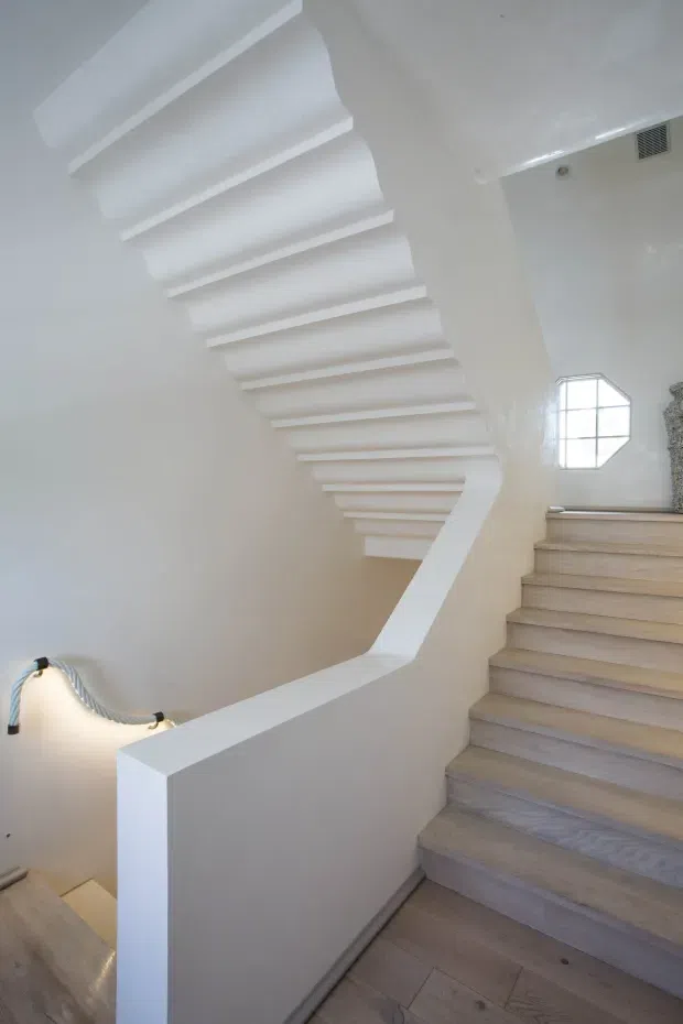 A bright, sculptural interior stairway with pale timber treads, white plaster walls, and a curved white balustrade. The staircase features a dramatic sculpted underside with repeated horizontal rib-like forms, a geometric window at the landing, and concealed accent lighting along the lower stairwell.