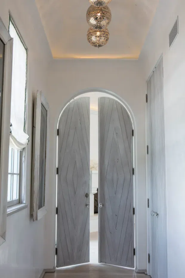 Bright, narrow residential hallway or entryway with white walls, a recessed tray ceiling, and three ornate metallic spherical pendant lights. The focal point is a pair of tall arched double doors in pale grey-washed timber with geometric plank detailing. A tall window with a light Roman blind is on the left, with additional timber-framed doors or shutters along the corridor. The space has a refined, decorative coastal-traditional feel.