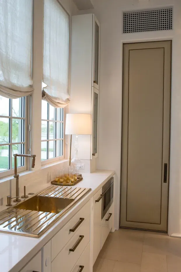 A narrow, bright butler-style kitchen or scullery with a long white stone benchtop, integrated cabinetry, a deep brass-toned apron sink with drainboards, tall multi-pane windows with fabric Roman blinds, and a concealed-panel door. Brass and dark metal hardware, a glass table lamp, and a small drinks tray add refined traditional detailing.