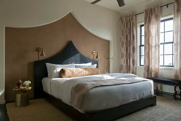 A refined bedroom with a large upholstered bed set against a dramatic curved, sculptural feature wall. The room combines warm taupe wall finishes, dark timber or painted bed joinery, textured neutral bedding, patterned full-height curtains, brass wall sconces, a woven carpet, and a black-framed multi-pane window. A ceiling fan and a small side table complete the space.