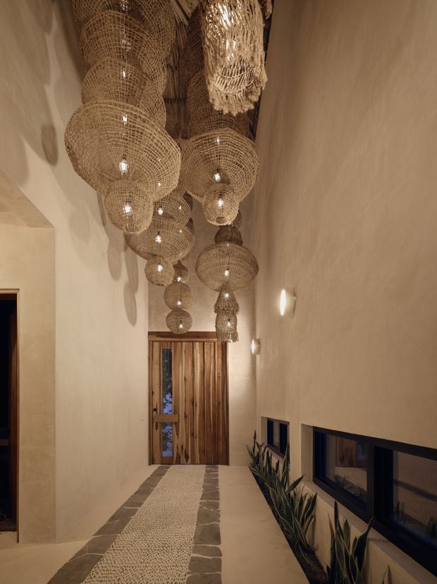 A tall, narrow residential hallway or entry passage with warm textured plaster walls, a timber front door, low horizontal windows, and a long runner bordered by irregular stone tiles. Numerous woven natural-fibre pendant lights hang in a dramatic clustered installation from the high ceiling, creating a warm, earthy, handcrafted atmosphere. Built-in planter beds with tall leafy plants line the window wall.