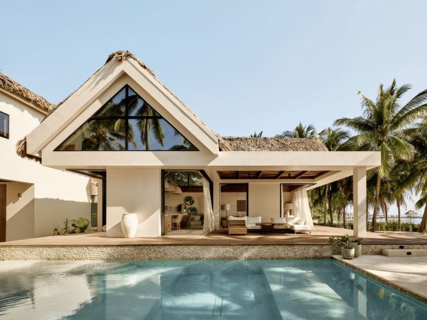 Exterior view of a tropical resort-style villa with cream rendered walls, steep triangular gable, expansive dark-framed glazing, thatched roof sections, and an open covered outdoor living area facing a swimming pool. Palm trees and bright coastal sunlight reinforce the tropical setting.
