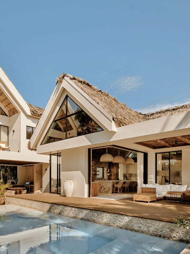 Exterior view of a tropical resort-style residence with white rendered walls, steep triangular gables, expansive black-framed glazing, and layered thatched roofing. The open-plan living and dining area extends onto a timber deck beside a swimming pool, with natural timber, woven pendant lights, and a stone-faced bar visible inside.