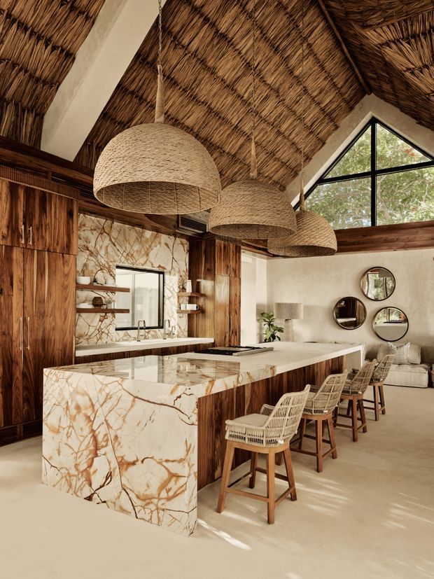 A warm, tropical-inspired open-plan kitchen and living area with a dramatic pitched ceiling lined in woven thatch or reed, exposed timber framing, and three large woven pendant lights. The kitchen features dark timber cabinetry, a pale marble or natural-stone island and benchtop with prominent brown veining, open shelving, and a black-framed triangular clerestory window. Woven bar stools line the island, while a small lounge area with a light sofa and round wall mirrors is visible beyond.