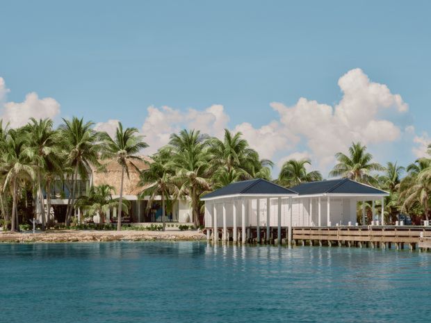 A tropical waterfront property viewed across clear blue water, featuring a low-rise pavilion or boathouse on stilts with a dark hipped roof, white posts and railings, and a timber waterfront deck. Behind it are palm trees and additional resort-style buildings with light walls and thatched or pitched roofs. The scene is sunny, relaxed, and distinctly coastal or island-oriented.