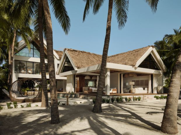 Exterior view of a low-rise tropical resort-style residence surrounded by tall palm trees. The building has multiple steep triangular gables, prominent light-coloured structural framing, open-air living areas, extensive glazing, and textured thatched roofs. Sandy ground, lush planting, and shaded outdoor seating reinforce the warm coastal setting.