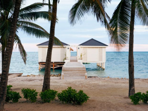 A tropical beachfront scene framed by palm trees, showing two small white waterside pavilion structures with dark pitched roofs, connected by a timber walkway extending over clear blue water. Sandy beach and low coastal planting occupy the foreground.