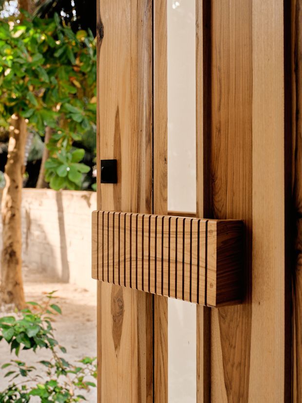 Close-up detail of a vertically timber-clad door or gate with a projecting slatted timber pull handle and a small black latch. The natural wood grain and warm tones dominate, while a leafy courtyard and pale masonry wall are visible beyond the opening.