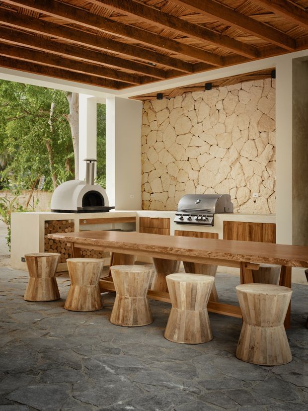 A covered outdoor entertaining area with a built-in kitchen, featuring a long solid timber dining table and rustic wooden stools in front of a stone feature wall. The cooking area includes a dome-shaped pizza oven, stainless steel barbecue, timber cabinetry, and open firewood storage. A richly textured exposed timber-beam ceiling and stone-paved floor create a warm, natural setting surrounded by greenery.