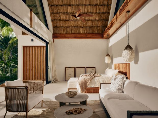 A bright, airy bedroom-lounge suite with a low-profile bed, white upholstered sofa, woven lounge chairs, layered round coffee tables, and neutral textured textiles. Large sliding glazing opens toward lush tropical greenery, while timber-framed detailing, a woven thatched ceiling, exposed beams, pendant lights, and a timber slatted screen create a warm resort-like atmosphere.