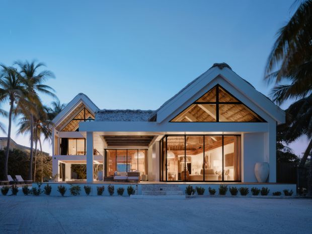 A tropical contemporary house viewed from the front at dusk, with white rendered walls, steep gabled roof forms, exposed timber detailing beneath the gables, and extensive floor-to-ceiling glazing. The open-plan interior is warmly illuminated, while palms and low landscaped planting frame the exterior. A covered outdoor living area connects directly to the glazed main living space.