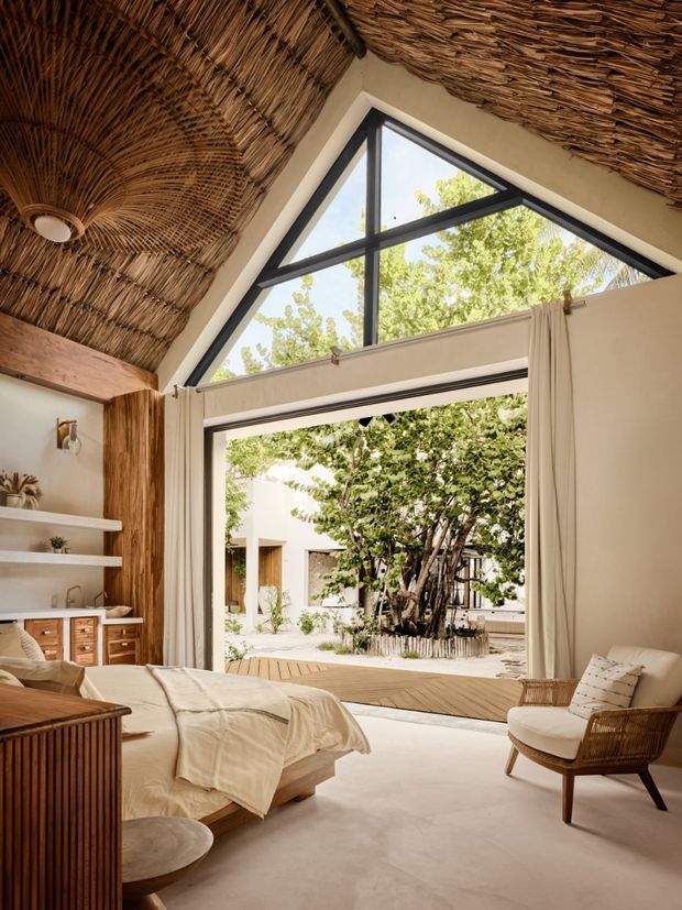 A warm, tropical-inspired bedroom with a low platform bed, woven rattan chair, timber furniture and exposed natural reed thatching across the vaulted ceiling. Large black-framed triangular clerestory glazing and wide open doors connect the room to a leafy internal courtyard. Neutral plaster walls, cream curtains and soft natural textiles create a relaxed resort-like atmosphere.