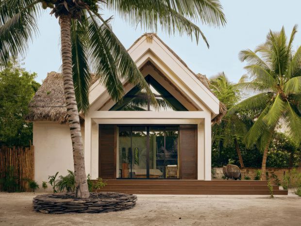 Front exterior view of a tropical, single-storey gable-roof pavilion or house surrounded by coconut palms. The pale rendered facade has a prominent triangular gable, deep dark-green recessed detailing, large glazed sliding doors, a timber-toned deck and a thatched side roof structure. The setting appears lush and resort-like, with sandy ground and tropical planting.