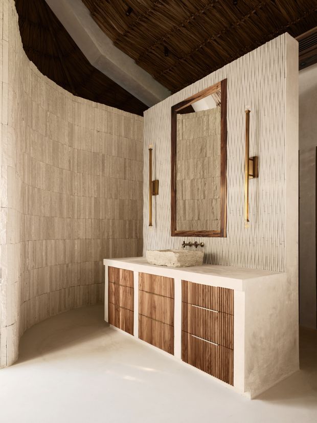 A warm, sculptural bathroom vanity area with a white textured built-in vanity, integrated stone basin, timber slatted cabinetry, dark timber-framed mirror, and slim brass wall sconces. The curved walls are clad in light textured tile or plaster, while the ceiling features exposed natural reed or thatch detailing, creating an organic, resort-like atmosphere.