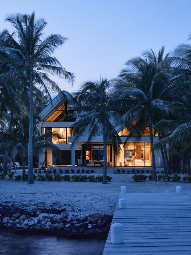 Twilight exterior view of a contemporary multi-level tropical residence, partially screened by tall palm trees. The home features extensive warm-lit glazing, dark structural framing, angular pitched roof forms, and a landscaped frontage opening toward a timber boardwalk and calm water in the foreground.