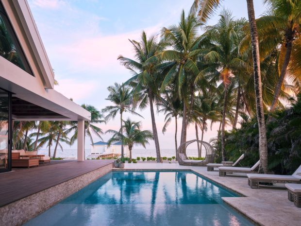 A tropical beachfront residence viewed from the pool, featuring a modern open-sided pavilion with broad roof overhangs, timber decking, a rectangular swimming pool, pale stone paving, lounge furniture, and dense coconut palms framing the ocean beyond. The scene has a relaxed, luxurious resort-like atmosphere at sunset.