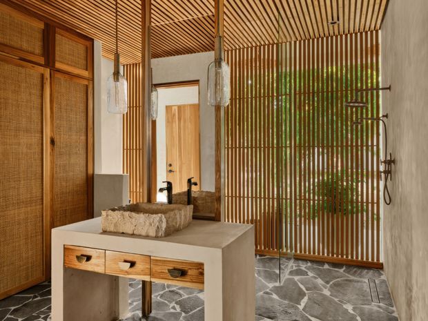 A warm, naturalistic bathroom with a freestanding stone vessel basin set on a minimalist concrete vanity with timber drawers. The space features extensive vertical and ceiling timber battens, woven cane wall panels, a timber door, exposed concrete surfaces, a frameless glass shower enclosure, and an irregular dark stone floor. Pendant lights and black tapware add subtle contrast, while greenery is visible beyond the screened opening.