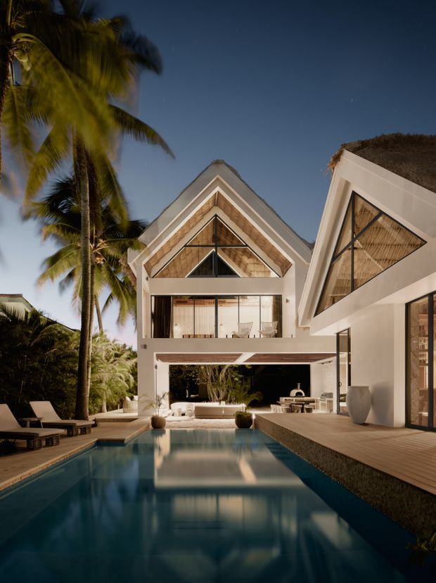 A twilight exterior view of a large tropical residence arranged around a long rectangular swimming pool. The white-rendered multi-level building has steep triangular gable forms, expansive dark-framed glazing, natural woven or timber-like triangular infill panels, and broad timber decking. Palm trees and poolside loungers create a resort-like setting, while warm interior and landscape lighting reflect across the water.