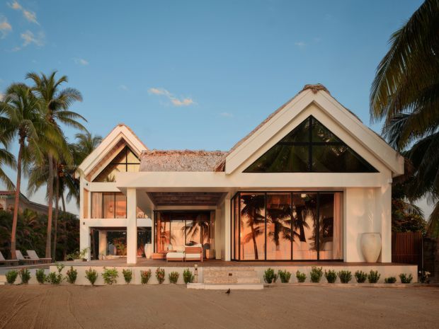 Exterior view of a tropical contemporary residence with prominent intersecting gabled roof forms, white rendered walls, broad glazed openings, timber-toned roof detailing, and a landscaped forecourt framed by palm trees.