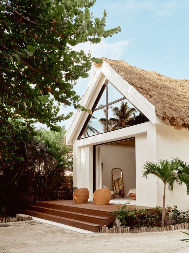 Exterior view of a tropical, resort-like gabled building with a steep thatched roof, white rendered walls, large triangular glazing, and wide open sliding doors. A timber deck with rounded wooden chairs leads into the bright interior, surrounded by lush palms and dense planting.