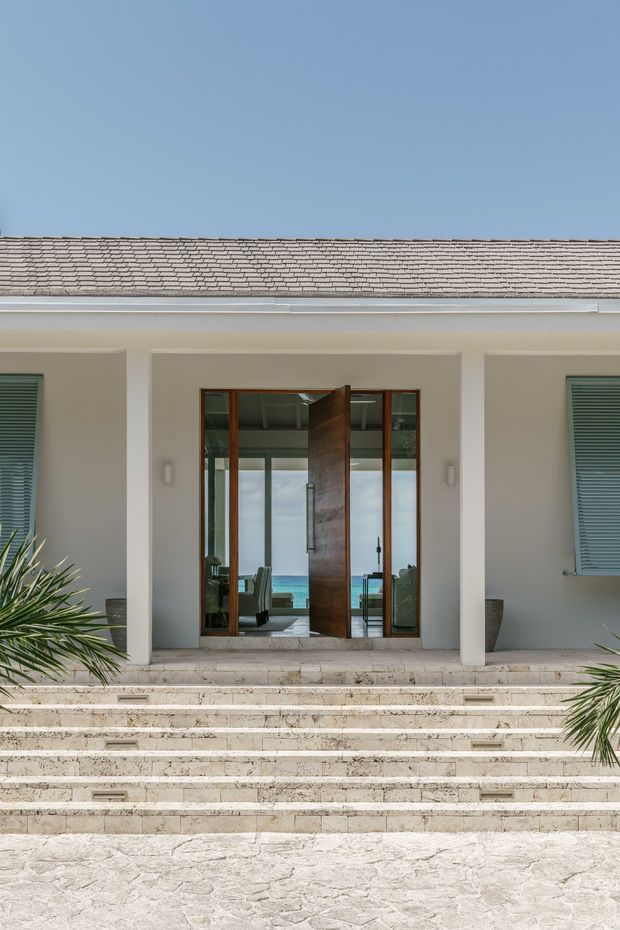 Front exterior entrance of a refined coastal-style residence, viewed from a broad pale stone staircase. The facade has smooth light-coloured rendered walls, deep rectangular entry recesses, timber-framed glazing, a large pivot timber door, louvred window shutters, and a low grey tiled roof. A glimpse of an interior living space and blue water is visible through the open doorway. Palm planting frames the steps.