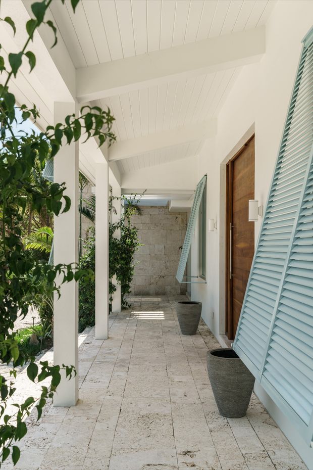 Bright covered exterior walkway or veranda with white square columns, a white-lined ceiling with exposed beams, pale stone or travertine paving, timber entry doors, and pale blue louvred shutters. Tropical planting borders the open side, while dark textured planters line the wall. The image has a breezy, refined coastal/tropical character.