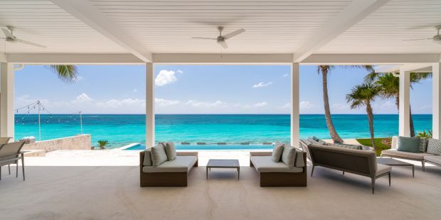 Open-sided covered outdoor lounge facing a vivid turquoise beachfront, with white structural posts, a slatted white ceiling, ceiling fans, and neutral upholstered seating arranged around a low coffee table. Palm trees and the ocean are visible beyond the terrace, creating a bright tropical coastal setting.