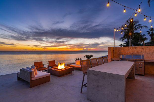 A beachfront outdoor entertaining terrace at sunset, with a concrete outdoor kitchen island and bar seating, built-in grill cabinetry, lounge seating, a fire pit, palm trees, and overhead festoon lighting overlooking calm water.