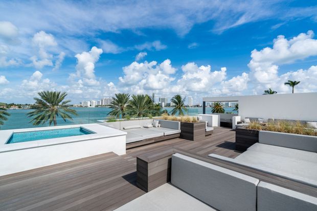 A bright modern rooftop terrace overlooking a waterfront, with a raised rectangular swimming pool, expansive timber decking, built-in white and grey upholstered lounge seating, and large integrated planters. The low white parapet and clean-lined architectural forms frame views of the water, palm trees, and distant high-rise buildings under a vivid blue sky.