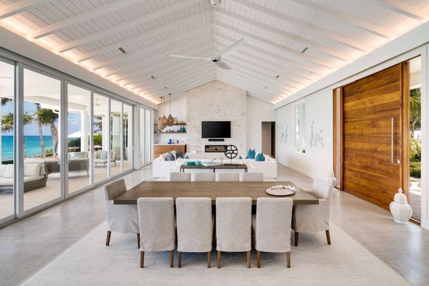 Bright open-plan coastal living and dining space with a long upholstered dining table in the foreground and a white sectional seating area beyond. The room features a white painted pitched, panelled ceiling with exposed-looking beams, integrated warm cove lighting, extensive floor-to-ceiling sliding glazing, polished concrete flooring, white walls, and a large natural timber pivot entry door. Tropical planting and a blue ocean view are visible outside through the glazing.