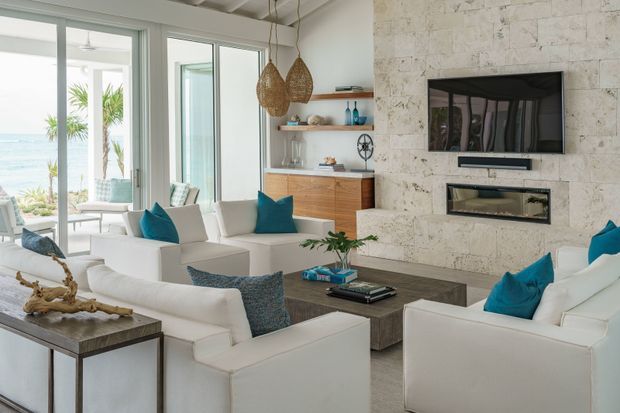 Bright coastal living room with white upholstered seating arranged around a dark rectangular coffee table, turquoise accent cushions, and a built-in media wall. The wall is clad in pale textured stone and incorporates a wall-mounted television above a linear fireplace. Floor-to-ceiling glazed doors open toward a sea view, while woven pendant lights, timber cabinetry, and open shelving add warmth.