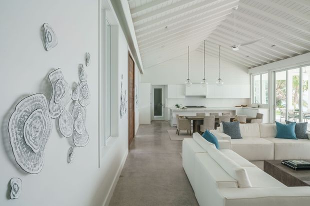 Bright open-plan living, dining and kitchen area with a large white sectional sofa in the foreground, a dining table and island beyond, and extensive windows along the right side. The room features a high white ribbed pitched ceiling with exposed beams, white walls and joinery, pale concrete or stone-look flooring, and minimalist organic wall artwork. Pendant lights hang above the dining and kitchen areas, while greenery is visible outside through the glazing.