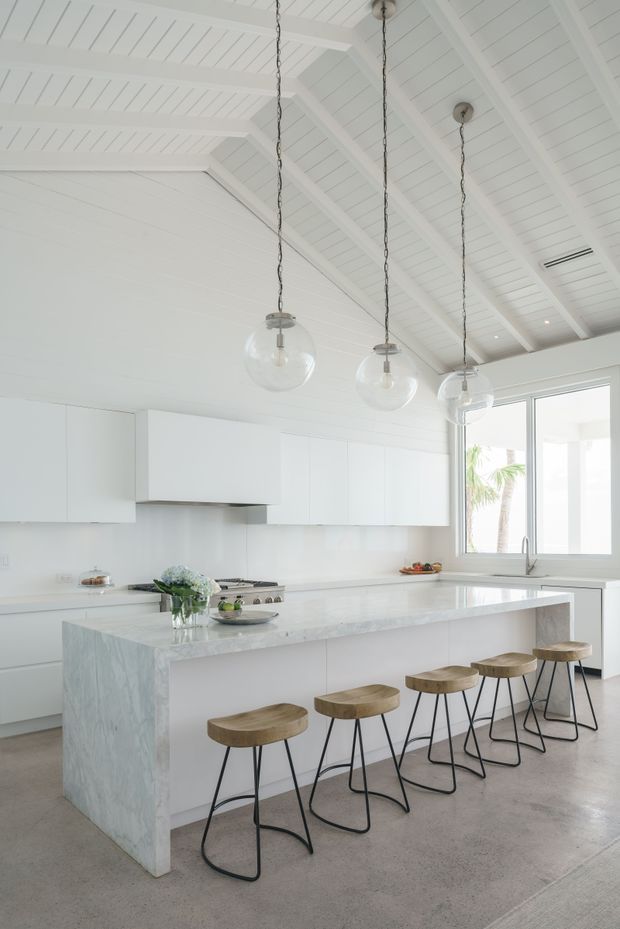 Bright, spacious white kitchen with a large waterfall-edge island, integrated white cabinetry, pale stone surfaces, polished concrete flooring, and four timber-and-black-metal stools. The room features a high vaulted ceiling clad in white boards with exposed white-painted beams, hanging globe pendant lights, and large windows providing abundant natural light.