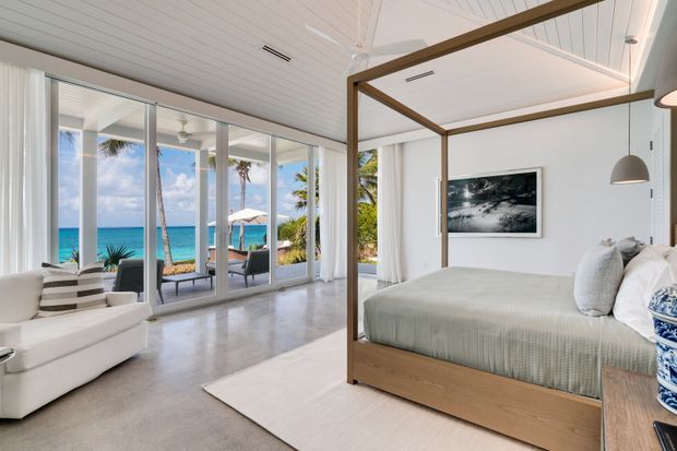 Bright coastal bedroom opening onto a tropical beachfront view. The room features a timber four-poster bed with pale bedding, white upholstered seating, full-height sliding glass doors, a white tongue-and-groove ceiling with ceiling fan, polished light concrete flooring, and sheer curtains. The ocean, palms, and outdoor seating are visible beyond the glazing.
