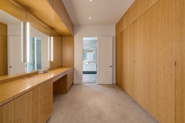 A warm, minimalist dressing room or walk-in wardrobe with extensive full-height timber cabinetry, a long built-in vanity with drawers, large mirror panels, and slim vertical pendant lights. The room has a pale textured floor, white ceiling and walls, and a central doorway leading to a bathroom or adjoining space.