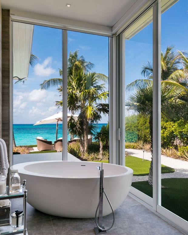 Bright contemporary bathroom featuring a freestanding oval bathtub positioned beside full-height corner glazing. The room overlooks a tropical landscaped garden and turquoise beach, with grey stone-look floor tiles, white walls, slim light-coloured window frames, and a minimalist chrome floor-mounted bath filler. A small glass-sided shelving unit and bathroom accessories sit at the left edge.