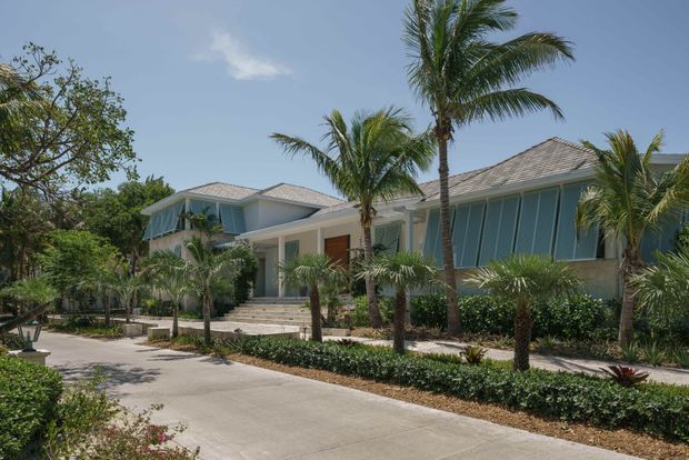 A low-rise tropical residential building viewed from a broad concrete driveway or walkway. The home has pale blue and white rendered walls, shallow hipped tiled roofs, generous glazed openings, covered entry verandas, and landscaped gardens with mature palm trees and clipped planting. The overall composition suggests a bright coastal or resort-like exterior.