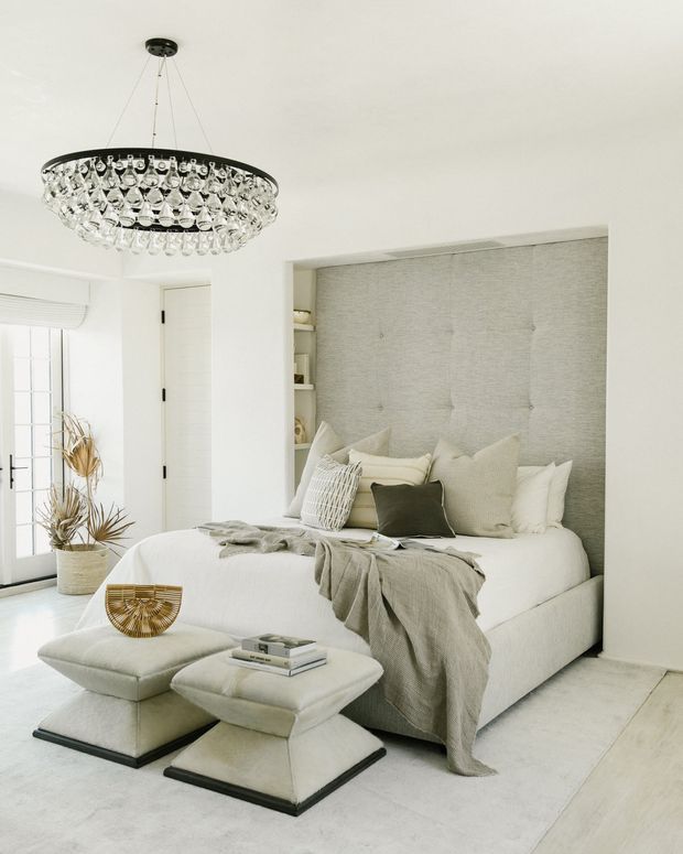 Bright, refined bedroom with a built-in upholstered bed set into a recessed alcove, textured light-grey wall panel, white bedding, layered neutral pillows, and a soft grey throw. Two upholstered ottomans sit at the foot of the bed, with a large crystal pendant light overhead. White walls, pale flooring, French doors, and restrained decor create an airy, luxurious neutral scheme.