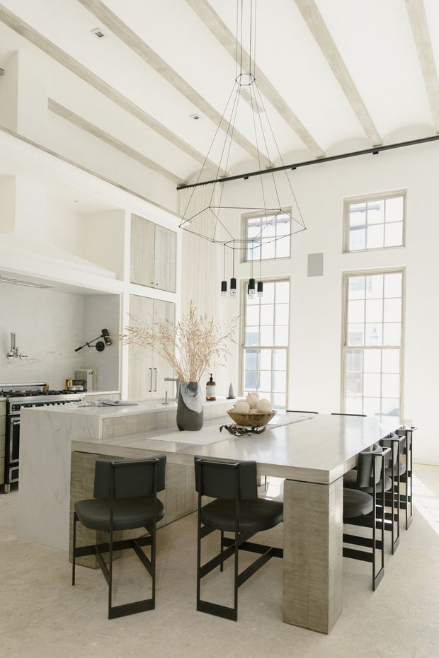 A bright, spacious open-plan kitchen and dining area with a large marble or pale stone island extending into a dining table, surrounded by dark upholstered bar stools and dining chairs. The room features white cabinetry, tall multi-pane windows, exposed pale ceiling beams, polished light flooring, and a dramatic geometric suspended pendant light. The overall design is refined, minimalist, and contemporary with strong natural light.