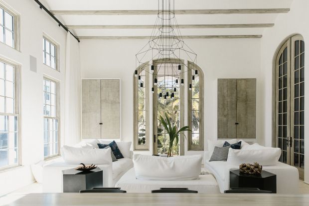 Bright, refined living room with white sofas arranged around dark square side tables, tall multi-pane windows and arched glazing, pale walls, exposed light-toned ceiling beams, neutral wall panels, and a large sculptural wire pendant. The room has abundant natural light and a high, elegant architectural character.