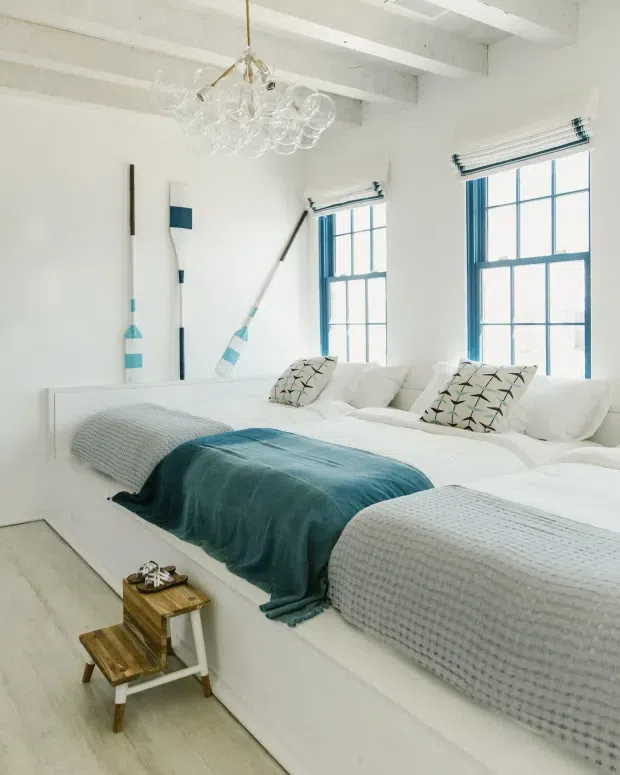 Bright coastal-style bedroom with two built-in white beds beneath exposed white ceiling beams. The room features white bedding, pale timber flooring, teal-blue throws and window frames, patterned cushions, decorative oars on the wall, and a cluster glass pendant light. Large blue-trimmed windows provide abundant natural light.