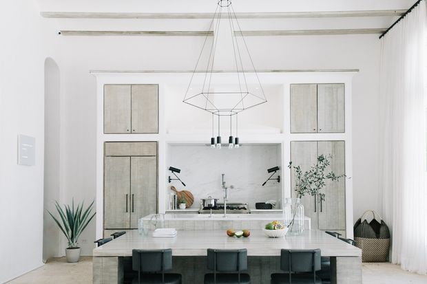 Bright, refined kitchen-dining space with a large central island and integrated dining seating. The room features white walls and cabinetry, pale timber or concrete-look cabinet fronts, a light stone island benchtop, marble-look splashback, exposed ceiling beams, tall sheer curtains, and a geometric suspended pendant light. Dark stools and black hardware provide restrained contrast.