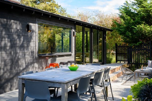 A contemporary single-storey backyard entertaining area beside a dark-clad pavilion or house extension. The image centres on a large outdoor dining table with modern chairs, set on a paved terrace next to floor-to-ceiling glazed doors and windows. Black vertical fencing, lush planted greenery, and a covered glazed room create a private garden setting. The low-sloping roof and horizontal dark cladding are clearly visible.