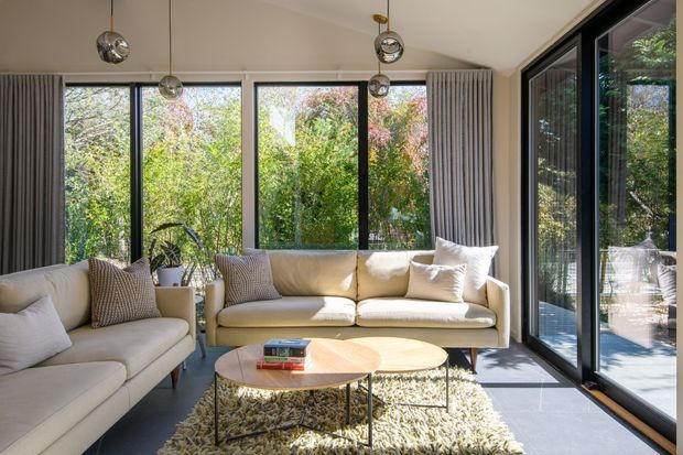 Bright contemporary living room with cream upholstered sofas, textured neutral cushions, layered round coffee tables, and a shag rug. Full-height black-framed glazing and sliding doors open the room visually to a leafy garden, while grey curtains and spherical pendant lights add contrast and texture.