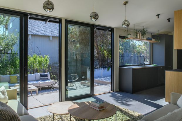 A bright contemporary open-plan living and kitchen area with extensive black-framed glazed doors opening to a landscaped patio. The room features dark kitchen cabinetry with timber accents, a pale ceiling, polished concrete flooring, a textured neutral rug, rounded timber coffee tables, and spherical pendant lights. Outdoor seating and greenery are visible beyond the glazing.