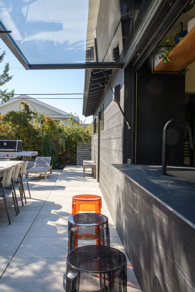 A sunny contemporary outdoor entertaining area beside a dark-clad house. The paved terrace includes a built-in outdoor kitchen or bar with a dark stone-like counter, black gooseneck tap, timber open shelving and a barbecue, along with stools, dining chairs and a table. A large glazed awning or roof panel extends overhead, and timber-like horizontal wall cladding and dark metal-framed openings define the house.